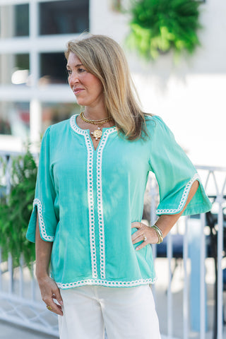 A medium close-up view highlighting the artisan detailing on the front of The Santorini Sun-Drenched Top. The shot showcases the rich turquoise mint fabric and the intricate, open-circle white geometric eyelet trim framing the neckline, center placket, and lower hemline. The model stands with one hand on her hip, accessorizing with a layered gold link necklace with a pearl-accented coin pendant and a stack of fine gold bracelets.