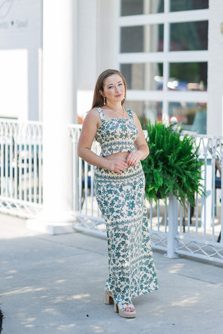Woman posing outdoors in a coordinated two-piece bohemian set featuring the Leya Tera Paisley Top and its matching maxi skirt. The cream smocked crop top showcases an intricate forest green floral and paisley block-print pattern, a clean square neckline, and a ruffled lettuce hem. Styled with a long gold link necklace, pearl drop earrings, and cream platform espadrille sandals.
