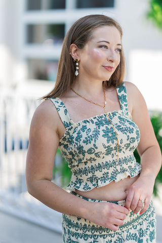 A medium close-up view highlighting the front details of the Leya Tera Paisley Top. The shot emphasizes the extensive, flexible smocking across the bodice and the crisp square neckline framed by wide, supportive shoulder straps. The fabric showcases a detailed green global-inspired botanical pattern against a cream woven background, accessorized with a gold lariat pearl necklace.