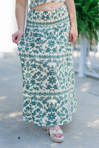 A close-up view highlighting the front design and textile detailing of the Leya Tera Paisley Maxi Skirt. The shot emphasizes the intricate green block-print design of mandalas, scrolling vines, and geometric borders against a cream woven viscose fabric. The model holds the edge of the flowing, straight skirt, showcasing the detailed stitching of the thick, stretchy smocked waistband.