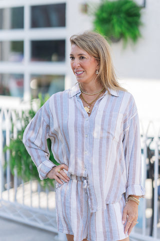 A medium close-up view highlighting the front design of the Z Supply Island Coconut Button Up. The shot showcases the multi-toned vertical earth-striped pattern in tan, blue, and cream on a soft linen-rayon fabric. Details include a classic pointed collar, a functional front button placket, and a single patch pocket on the left chest. The model poses with one hand on her hip, showing her rolled-up cuffs.