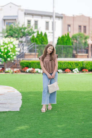 Full-length front view of a model posing on an outdoor green lawn showcasing the Desert Willow Lace Top. The loose, flowy brown top is paired with high-waisted, raw-hem wide-leg jeans and neutral double-strap block-heel platform sandals. She holds a cream woven purse, presenting a warm and modern bohemian boutique style.