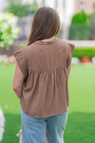 Rear view of a model from the waist up wearing the earthy brown Desert Willow Lace Top. The image highlights the structured drop-shoulder design that creates a cap-sleeve effect, as well as the curved back yoke seam with delicate gathering that opens into a relaxed, flowy lower bodice. Styled casually with light-wash blue jeans.