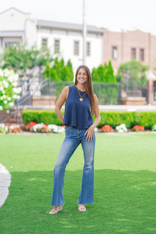 Full-length front view of a model standing on a manicured green lawn showcasing the Harbor Breeze Pleated Tank. The A-line swing top is paired with high-waisted flare jeans featuring raw frayed hems and neutral double-strap wedge sandals, presenting an elongated, casual boutique look in front of a soft-focus building.