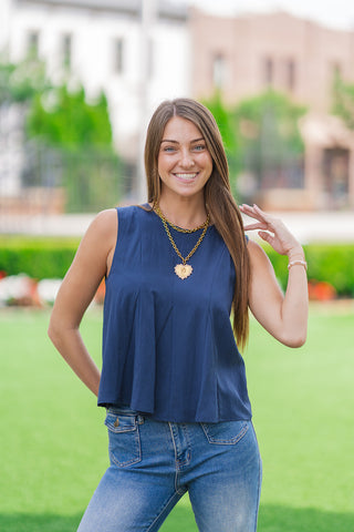 A front medium shot focusing on the tailored elements of the Harbor Breeze Pleated Tank. The dark blue sleeveless blouse showcases structured, stitched pleats fanning out down the front bodice to create a polished swing shape. The model smiles, accessorized with stacked gold link chains and a large heart pendant.