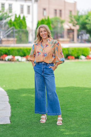 Full-length front shot of a model posing on a manicured lawn wearing the terracotta orange variant of the Whispering Winds Blouse. The relaxed-fit top is tucked into high-waisted wide-leg jeans, showcasing its airy silhouette and dramatic 3/4 balloon sleeves. The look is styled with stacked neutral block-heel slide sandals.