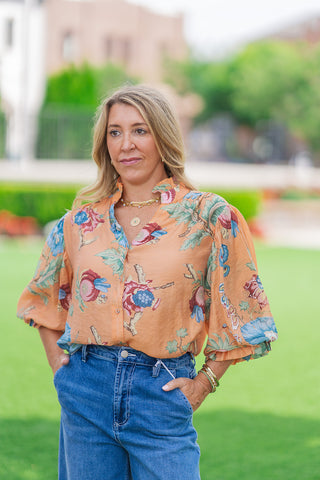 A medium front shot of a model wearing the terracotta orange option of the Whispering Winds Blouse. The image focuses on the button-down front with a ruffled stand collar, slightly opened into a V-neckline. The blouse features bold tropical blooms in blue and pink, paired with gold layered necklaces and bracelets.
