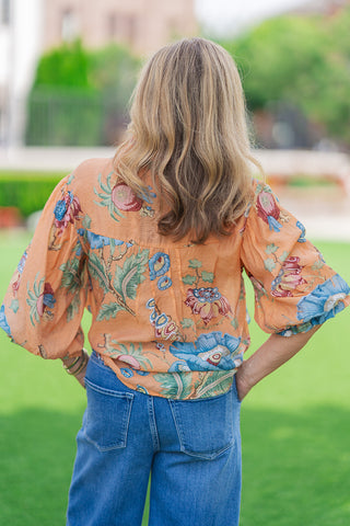 Rear view of a blonde model standing on a green lawn wearing the Whispering Winds Blouse in terracotta orange. The back panel highlights the lightweight woven fabric gathered subtly along the yoke, with voluminous 3/4-length balloon sleeves showing oversized blue, green, and pink botanical prints. Styled tucked into medium-wash wide-leg denim jeans.