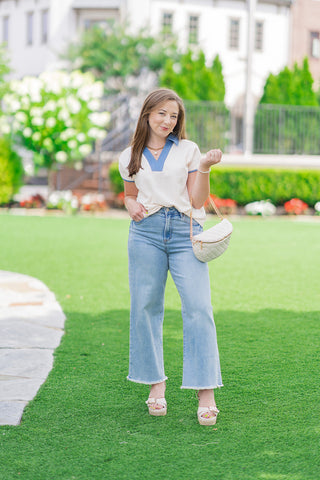 Full-length front view of a model posing on an outdoor lawn wearing the Seaside Chambray Top. The relaxed-fit cream blouse features a deep blue chambray V-neckline border and collar. It is styled for a casual boutique look with high-waisted wide-leg light-wash jeans, neutral woven platform sandals, and a quilted white crossbody belt bag with a gold chain strap.