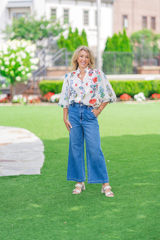 Full-length front view of a model showcasing the white option of the Whispering Winds Blouse. The top is casually styled with high-waisted, medium-wash wide-leg jeans and neutral double-strap block heels, emphasizing a modern bohemian aesthetic on an outdoor green lawn.
