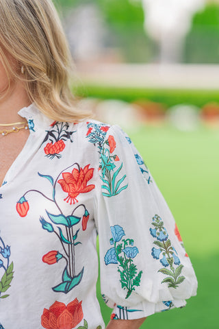 A close-up detail shot of the white floral Whispering Winds Blouse. The image focuses on the gathered shoulder seam that releases into a voluminous balloon sleeve, highlighting the crisp print layout of red, blue, and green bohemian botanicals. A delicate gold chain necklace is visible along the edge of the neckline.