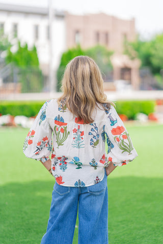 Rear view of a model from the waist up wearing the Whispering Winds Blouse in white. The shot emphasizes the structured back yoke and the vertically aligned floral motifs featuring orange tulips, green stems, and blue blossoms. The lightweight top drapes over high-waisted blue jeans against a soft-focus background.