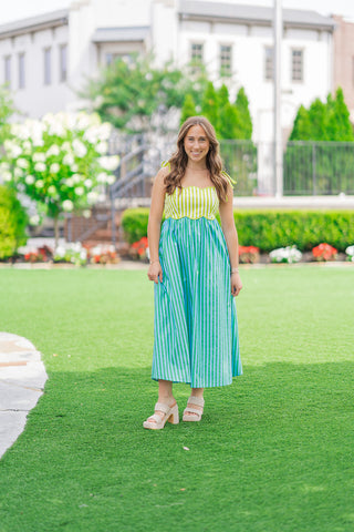 Full-length front view of a model posing on a manicured green lawn wearing the Key Lime Pop Stripe Dress. The sleeveless midi dress features a structured lime-and-white striped bodice contrasting with a lime-and-sky-blue striped, flowy skirt. She is smiling, styled for an elevated summer look with chunky neutral woven platform heels, with a white boutique building in the soft-focus background.