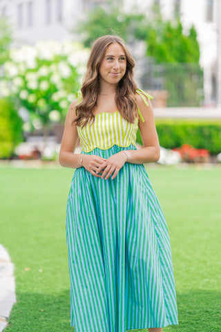 A portrait view of the Key Lime Pop Stripe Dress highlighting the vibrant lime green and white vertical-striped bodice. The close-up showcases the feminine contoured scallop neckline and the matching scalloped detail at the waistline. The model has her hands gently clasped, showing off the functional, wide shoulder tie straps against a blurred outdoor background.