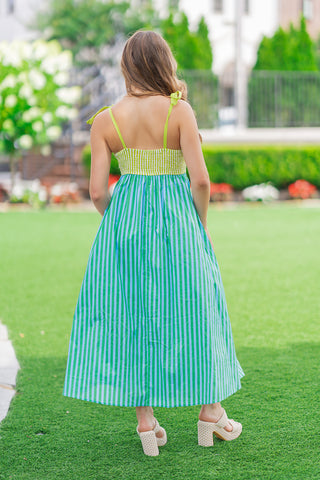 Back view of a model standing on a green lawn wearing the Key Lime Pop Stripe Dress. The photo highlights the flexible, lime-green smocked back bodice and the wide adjustable tie straps knotted at the shoulders. The flowy, vertical-striped midi skirt cascades down in alternating shades of lime green and sky blue, styled with neutral woven block-heel sandals.