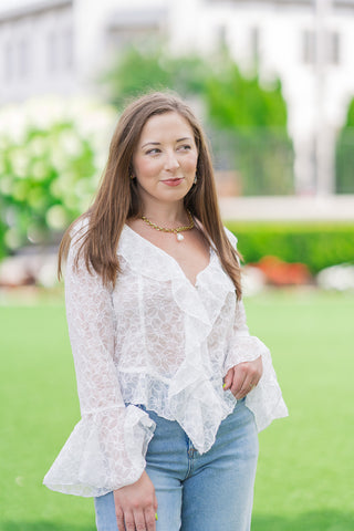 A front medium shot of a model standing outdoors wearing the white Fountain of Florals Lace Top. The sheer blouse showcases its romantic V-neckline framed by a cascading ruffled placket, structured tailored seams, and statement flared bell cuffs. The model is accessorized with a thick gold link chain necklace featuring a pearl pendant.