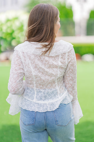 Rear view from the waist up of a model wearing the white Fountain of Florals Lace Top. The shot highlights the architectural paneling of the sheer floral lace, featuring structural vertical darts that taper down to a subtly flared peplum-style hemline. The top is styled casually with light-wash denim jeans against a soft-focus garden background.