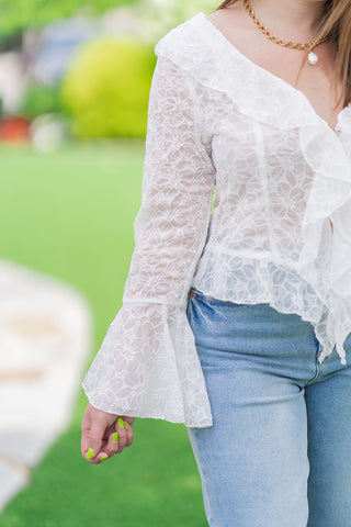 Close-up side profile view highlighting the intricate construction of the Fountain of Florals Lace Top. The image details the sheer, premium floral lace fabric, a structured vertical bodice seam, and the cascading ruffle trim along the V-neckline. The long sleeve finishes in a dramatic, flared tiered bell cuff over high-waisted light-wash blue jeans.