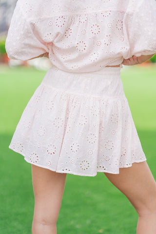 A rear close-up view from the waist down of a model standing on a green lawn wearing the light pink Rosewater Romance Tiered Skort. The image focuses on the thick, stretchy smocked elastic waistband and the breezy flow of the tiered, ruffled mini skirt fabric covered in allover floral eyelet cutouts.