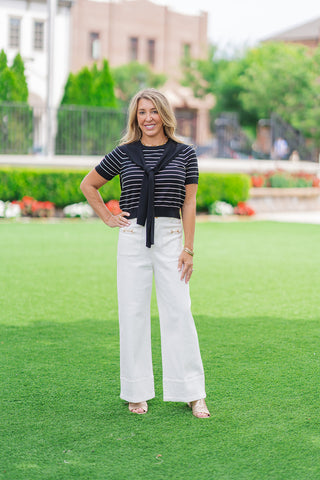 Full-length front view of a model posing on an outdoor lawn showcasing the Gold Standard Wide Leg Pants. The high-waisted white trousers feature crisp vertical seams down each leg and a wide-leg cut. The look is styled for a maritime aesthetic with a short-sleeve black striped knit top, an attached knit scarf tied at the chest, and gold slide sandals.