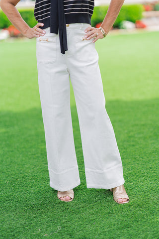 A front close-up shot from the waist down emphasizing the designer elements of the white Gold Standard Wide Leg Pants. The shot focuses on the high-waisted fit, front zip fly, and two prominent front patch pockets adorned with gleaming gold horsebit hardware accents. Sharp vertical seams line the center of each leg, styled with textured gold slide sandals.
