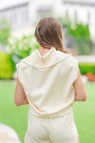 Rear view from the thighs up of a model posing on an outdoor lawn wearing the two-piece Sunshine Sorbet Gingham Set. The image emphasizes the wide, structured fold collar of the sleeveless top draping elegantly over the upper back, creating a sporty yet casual look in a yellow and white gingham pattern.