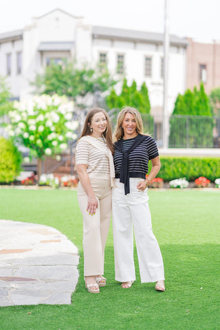 Two models stand together on a manicured green lawn showcasing both color options of the Parisian Port Stripe Top. The model on the left wears the ecru version with matching cream wide-leg pants and woven platform heels, while the model on the right wears the black version with white trousers and gold slides.