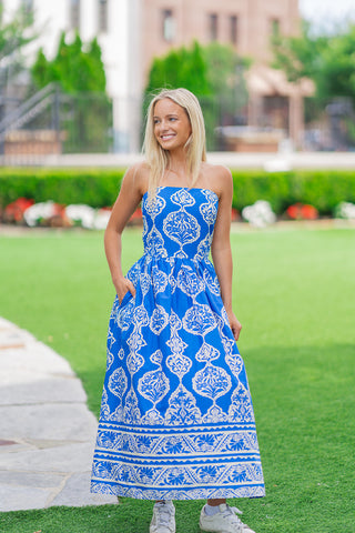 A medium front shot of the Aegean Sea Damask Midi Dress, showcasing the elegant strapless straight-across neckline. The model smiles and looks away, keeping one hand tucked into the functional, hidden side pocket. The deep ocean blue and crisp white damask pattern creates a flattering, structured look across the fitted bodice and flowing skirt.