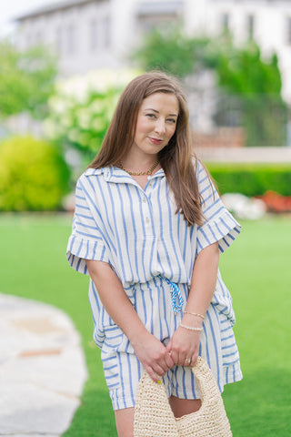 A front medium shot of a model highlighting the full Saint-Tropez Stripe Gauze set. The oversized, vertical-striped short-sleeve top is worn over the matching high-waisted shorts, revealing a glimpse of the blue and white twisted drawstring at the waist. The model holds a textured cream straw bag, styled with a gold link chain necklace.