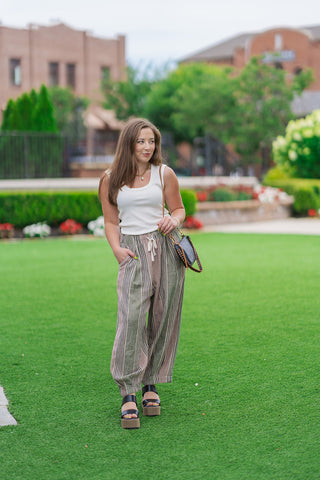 Full-length front view of a model posing on a manicured green lawn wearing the Meadowland Linen Pants. The ultra-relaxed, multi-colored vertical striped trousers are paired with a fitted white scoop-neck tank top and a black leather crossbody bag with a braided strap, creating an effortless bohemian boutique style.