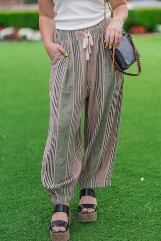 A front close-up shot from the waist down focusing on the relaxed construction of the Meadowland Linen Pants. The image highlights the gathered elastic waistband with its functional cream drawstring tie, deep side pockets, and the loose balloon leg silhouette that tapers gently toward the ankle. The model stands on grass wearing chunky black double-strap platform wedges.
