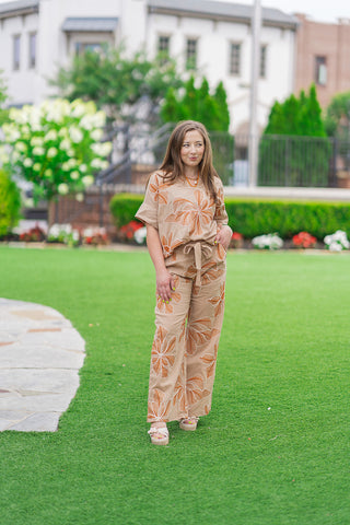 Full-length front view of a model posing on an outdoor green lawn showcasing the Sahara Sunflower Pants. The high-waisted, wide-leg beige trousers are paired as a matching two-piece coordinate set with a short-sleeve popover blouse, styled with neutral platform sandals for a sophisticated summer resort aesthetic.