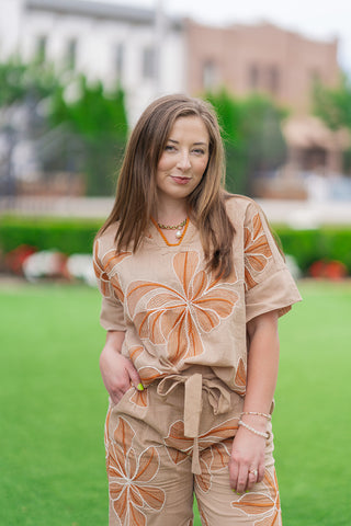 A front medium shot highlighting the artisan features of the Sahara Sunflower Top. The beige popover blouse showcases oversized, rust-and-white embroidered floral motifs across the front bodice and sleeves. The open split V-neckline is trimmed with coordinating mustard-orange mini pom poms, accessorized with a gold pearl pendant necklace.