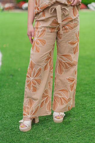 A front close-up shot from the waist down focusing on the artisan details of the Sahara Sunflower Pants. The high-waisted beige trousers highlight a smooth front waistband with an attached fabric tie belt and deep side pockets. The flowing wide legs showcase the high-contrast white stitched outlines of the embroidered sunflowers, styled with double-strap platform wedges.