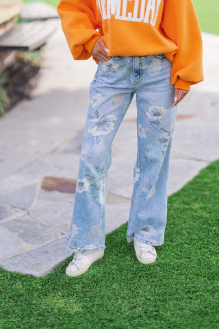A model showcases the English Garden Rigid Flares, featuring a muted pastel floral print over light-wash blue denim. The high-quality non-stretch jeans are styled with an oversized, vibrant orange crewneck sweatshirt and white low-top sneakers. The photo captures a front view of the structured wide-leg silhouette against a backdrop of a stone walkway and green grass.