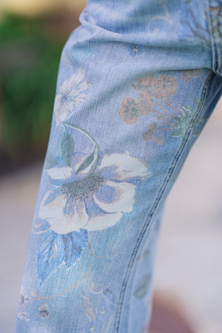 A detailed close-up shot of the English Garden Rigid Flares, highlighting the premium rigid denim fabric texture. The image showcases the intricate allover botanical pattern, featuring soft cream-colored blossoms, delicate green leaves, and muted tan stems seamlessly overlaid on the light-blue wash base denim.