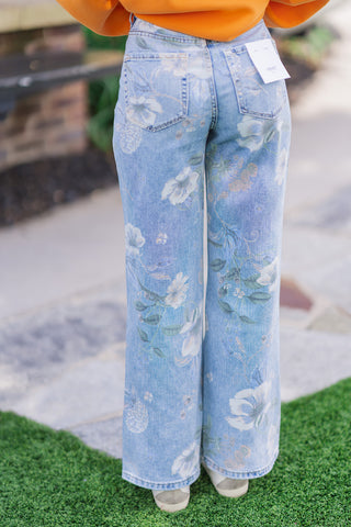 Back view of the English Garden Rigid Flares, emphasizing the traditional 5-pocket construction and clean, finished hemline. The model stands on green grass wearing the high-waisted, retro-cut flare jeans with a white Vervet brand tag attached to the back right pocket. The oversized orange sweatshirt is slightly cropped at the back, showcasing the flattering, structured fit.