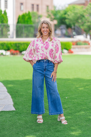 Full-length front view of a model posing on an outdoor green lawn showcasing the Versailles Paisley Blouse. The lightweight woven top with dramatic 3/4 balloon sleeves is tucked into high-waisted wide-leg denim jeans and styled with neutral double-strap block heel sandals, creating an elegant summer boutique look.