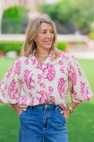 A front medium shot highlighting the intricate details of the Versailles Paisley Blouse. The cream button-down top features an elaborate magenta floral print, an open split V-neckline, and a delicate ruffled stand collar. The model stands with hands in her pockets, accessorized with layered gold necklaces and stacked bracelets.