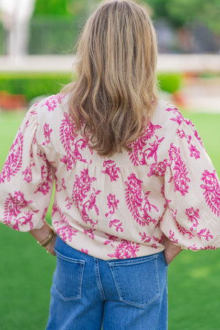 Rear view of a model from the waist up wearing the Versailles Paisley Blouse. The image focuses on the cream-colored fabric detailed with a vivid magenta-pink paisley and floral print, featuring a clean back yoke with subtle gathering that flows into the relaxed bodice and voluminous 3/4-length balloon sleeves. Styled tucked into medium-wash blue jeans.