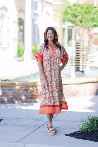 Woman smiling outdoors in the Aperol Spritz Midi Dress, a flowy kaftan-style piece featuring an intricate, tapestry-inspired botanical floral pattern in earthy terracotta, peach, and gold tones. The dress is framed by high-contrast tangerine and poppy red color-blocked panels at the split v-neckline, wide dolman sleeves, and sweeping border hemline. Styled for a vibrant, effortless summer resort look with tan platform espadrille sandals.