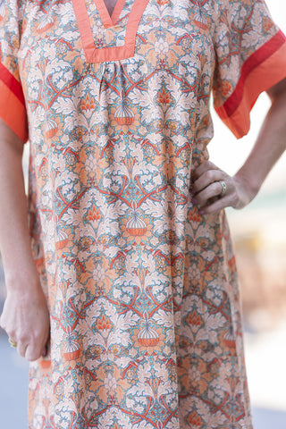 A close-up detail shot focusing on the bodice of the Aperol Spritz Midi Dress. The image perfectly captures the lightweight, unlined woven fabric and the ornate, vintage-inspired mix-floral pattern. The vibrant tangerine contrast binding at the split v-neckline stands out beautifully against the warm, earthy tones of the botanical print, showcasing the garment's high-end boutique quality.