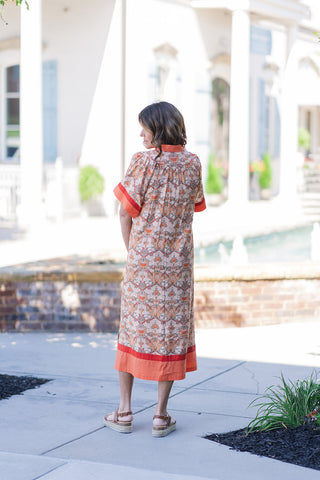 Back view of the Aperol Spritz Midi Dress, showcasing the forgiving, fluid fit of the relaxed kaftan silhouette. The image details the continuous floral tapestry pattern and the sweeping straight midi hemline, which features a bold contrast border and dynamic side slits that allow for breezy movement. The model is standing outdoors in tan platform espadrilles, looking gracefully over her shoulder.