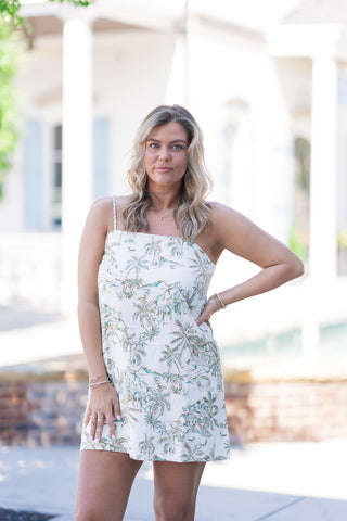A closer front view of a woman wearing the Z Supply Maude Tiki Tide Mini Dress, highlighting the comfortable shift-style silhouette that skims beautifully away from the torso. The premium washed linen-rayon fabric drapes fluidly, showcasing the whimsical toile-like tropical coastal print. The model rests her hand casually on her hip, accessorized with layered gold bracelets and rings.