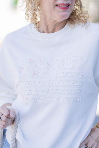 A close-up front view of a woman wearing the Sweet Land of Liberty Sweatshirt, highlighting the unique heirloom aesthetic of the cottagecore-inspired American flag applique. The Canton star block features a vintage-inspired pink rose print, while the stripes alternate with coordinating calico florals in muted pink, sage, and dusty blue hues. The image details the raw-edge tactile texture of the stitched fabric panels and the classic ribbed knit crew neck against the ultra-soft white fleece pullover.