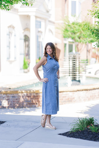 Full-length outdoor portrait of a smiling woman posing to demonstrate the fluid movement of the Dear John Luella Midi Dress. The sleeveless silhouette is structured with a defined waist seam and cascades into an A-line skirt with a clean, wide-topstitched hem. She styles the casual washed-denim dress for a chic daytime look with metallic espadrille wedge sandals, set against a blurred background with a water fountain.