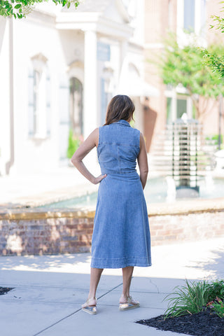 Back view of a woman showcasing the clean, structured silhouette of the Dear John Luella Midi Dress. This sleeveless denim piece features a defined waist seam, a straight back yoke line with vertical princess seams for a tailored fit, and a wide-stitched hemline resting at a midi-to-maxi length. The model is turned away, accessorized with metallic wedge sandals against a sunny resort-style courtyard backdrop.