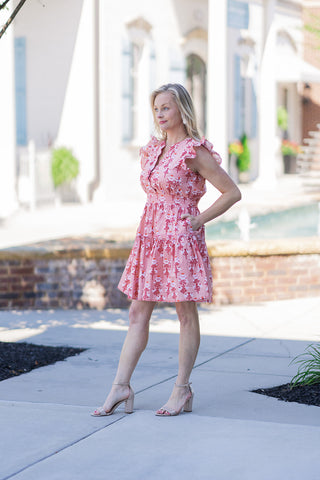 Woman posing outdoors in the Peachy Keen Ruffle Mini Dress, a flirty and youthful piece featuring a vintage-inspired floral print in soft peach and warm coral tones. The dress has airy flutter ruffle short sleeves, a comfortable cinched waist, and a flowing mini-length skirt with functional side pockets. Styled for a picture-perfect daytime look with nude block-heeled sandals.