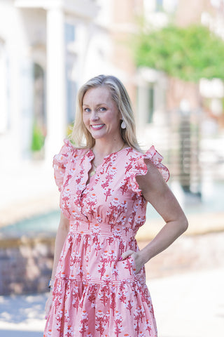 A smiling portrait view highlighting the bodice of the Peachy Keen Ruffle Mini Dress. The image showcases the unique split v-neckline secured by an intricate, scalloped snap-closure trim. The lightweight, fully lined woven fabric displays a delicate allover botanical pattern, framed by feminine flutter sleeves. The model rests one hand casually in her pocket, accessorized with white drop earrings and a subtle gold pendant.