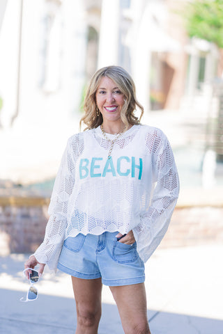 A portrait view highlighting the Saltwater Breeze Sweater's intricate white chevron-inspired pointelle knit and delicate scalloped edge hemline. The woman smiles warmly, posing with one hand resting in the pocket of her light-wash denim shorts, perfectly showcasing the bold teal "BEACH" intarsia lettering, layered gold necklaces, and the flowy drape of the bohemian bell sleeves.